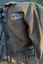 Load image into Gallery viewer, A brown canvas jacket with patch style side pockets and an upper left chest zipper pocket with a brand patch above.